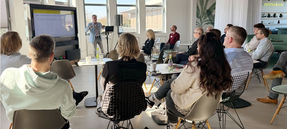 Eine Gruppe von Menschen sitzt in einem modernen, hellen Raum und hört einer präsentierenden Person zu, die vorne neben einem großen Bildschirm steht. Auf Tischen stehen Getränke, Notizmaterial und Laptops. Die Teilnehmenden sind in einer halbkreisförmigen Sitzordnung angeordnet und folgen aufmerksam der Präsentation.