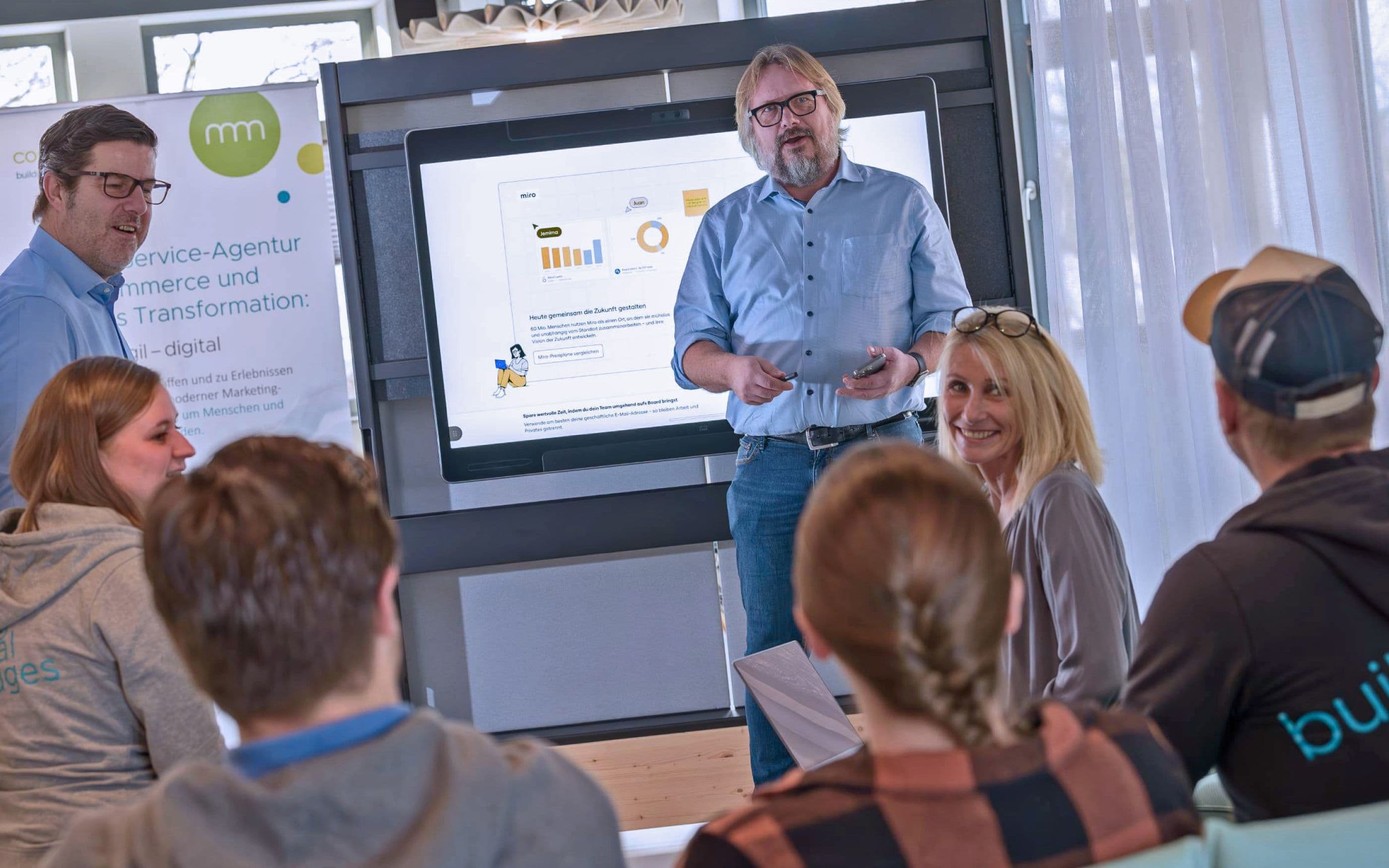 Eine Gruppe nimmt an einem Workshop teil, während ein Vortragender vor einem großen interaktiven Bildschirm steht und Diagramme sowie Inhalte präsentiert. Die Teilnehmenden sitzen im Raum und folgen der Präsentation in einer modernen Arbeitsumgebung.